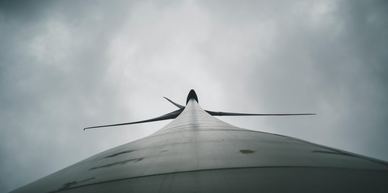 Widok z dołu na wysoką turbinę wiatrową z rozłożonymi łopatami na tle pochmurnego nieba.