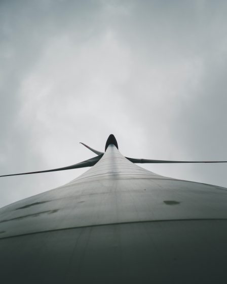 Widok z dołu na wysoką turbinę wiatrową z rozłożonymi łopatami na tle pochmurnego nieba.