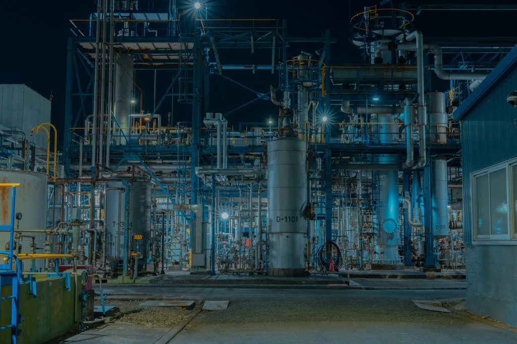 Oświetlone instalacje przemysłowe z rurami, zbiornikami i konstrukcjami stalowymi w dużym zakładzie produkcyjnym nocą.
