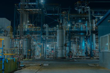 Oświetlone instalacje przemysłowe z rurami, zbiornikami i konstrukcjami stalowymi w dużym zakładzie produkcyjnym nocą.