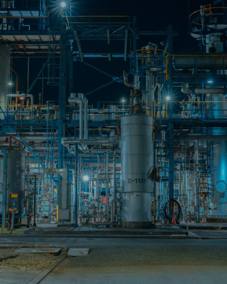 Oświetlone instalacje przemysłowe z rurami, zbiornikami i konstrukcjami stalowymi w dużym zakładzie produkcyjnym nocą.