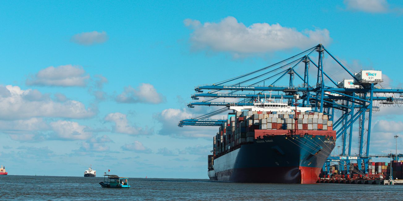 Duży statek kontenerowy zacumowany w porcie przy suwnicach przeładunkowych i stosach kontenerów.