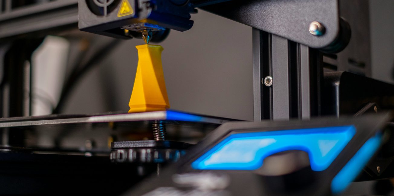 Głowica drukarki 3D tworząca żółty element z filamentu na platformie roboczej urządzenia.