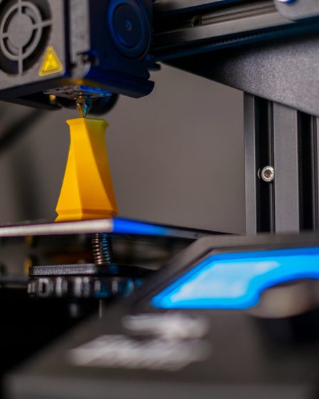 Głowica drukarki 3D tworząca żółty element z filamentu na platformie roboczej urządzenia.