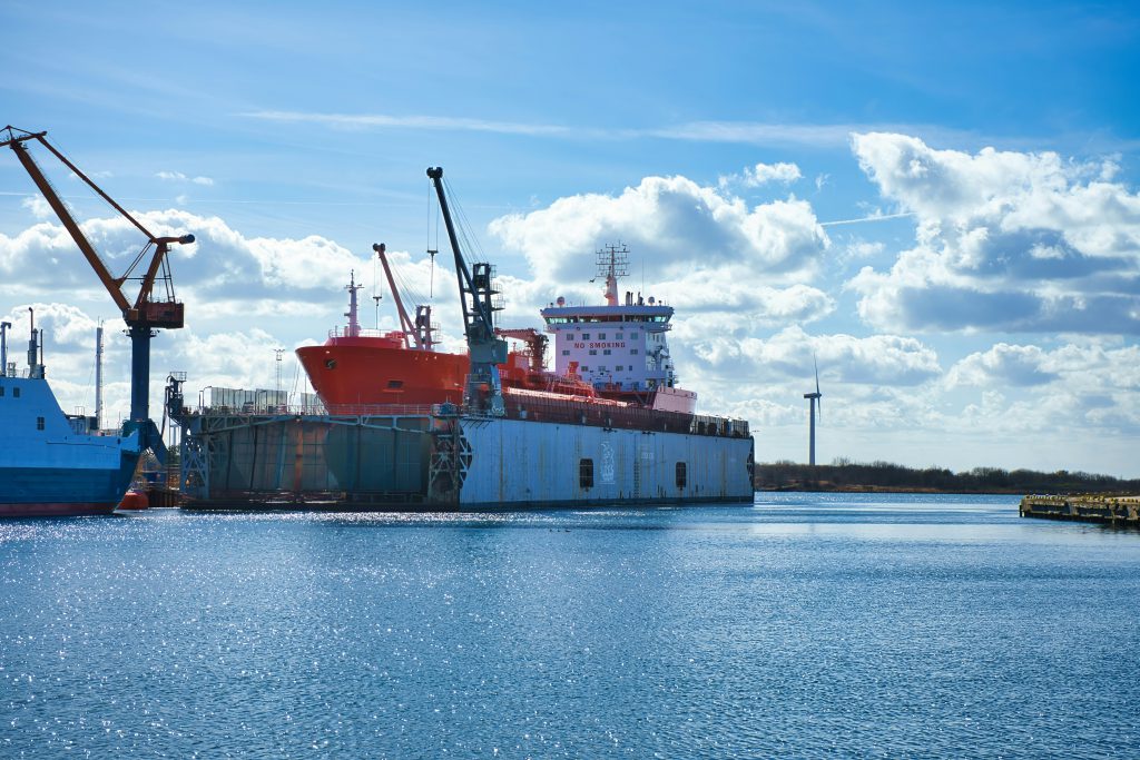 Statek towarowy w porcie morskim z dźwigami portowymi oraz instalacjami przemysłowymi nad wodą.